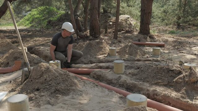 A man in a hard hat installs sewage pipes in a forested area.