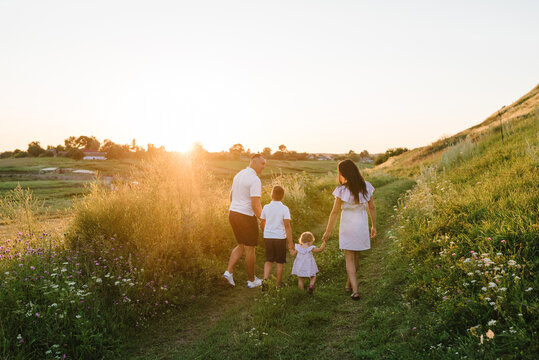 A mother, father holding hands baby daughter, son walk in mountains at sunset. Family spending time together on summer day. Children with parents happiness walking in green grass in field. Back view