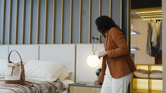 African American businesswoman preparing for work, adorning with earrings in a well-appointed bedroom. Poised and ready, woman grabs stylish purse before leaving home for the day