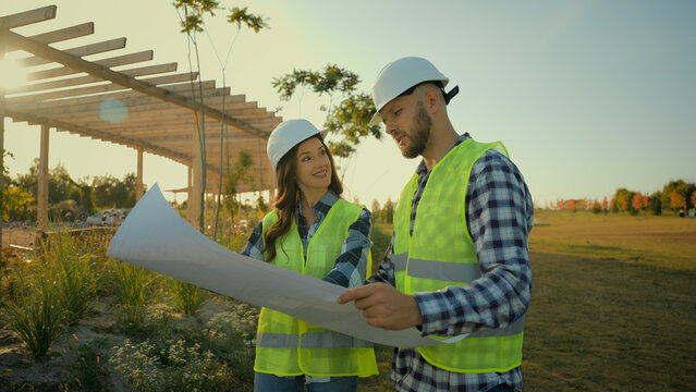 Two Caucasian happy professional engineers smiling successful builders contractor woman female girl man guy helmet male workers talking discuss holding blueprint plan outside farm business field park