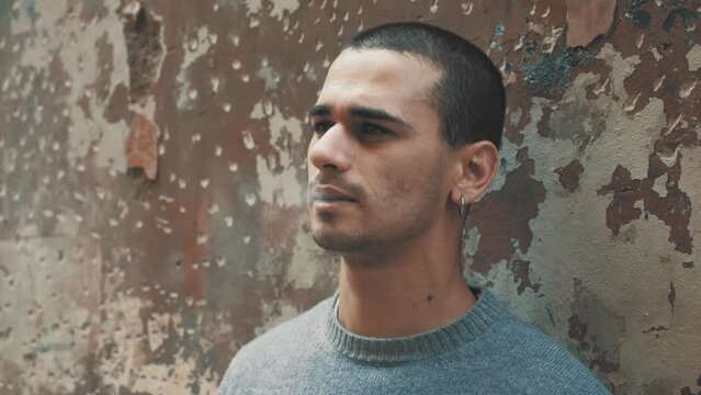20-year-old man with short hair in gray sweater, leaning against wall in a suburban courtyard, smoking a cigarette with a very serious expression.