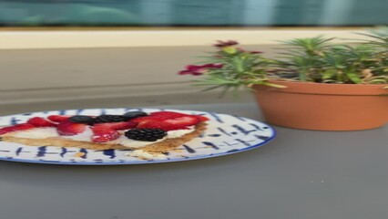 A close-up view of a tart with cream, strawberries, blackberries and blueberries. Vertical video of a pie with berries and a pot of flowers on a table in a cafe - Powered by Adobe