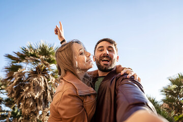 Happy couple taking a joyful selfie outdoors with palm trees and a blue sky. They are smiling and...