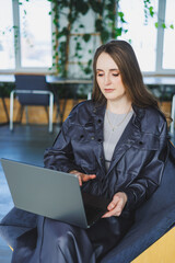 A young woman in a leather jacket sits in a cafe and works on a laptop. Beautiful girl in casual clothes.