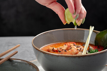 A popular dish of Asian cuisine is Tom Yam soup, cooked with shrimp, lemongrass, galangal and lime. A hand squeezes a drop of lime into a Thai soup with a sour-spicy taste.