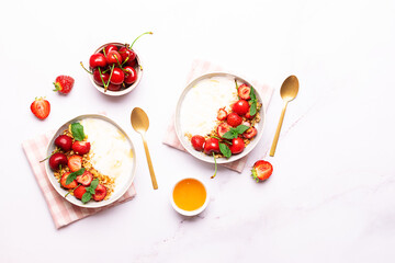 Greek yogurt with granola and fresh strawberries, cherry and honey, decorated with aromatic mint leaves on white marble background top view. Healthy tasty homemade breakfast concept. 