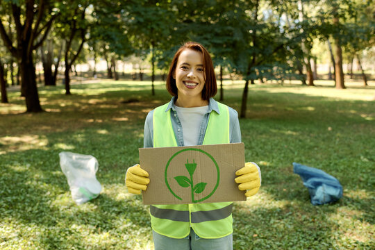 A woman in a green vest holds a cardboard sign, her expression reflective of a plea for help or awareness.