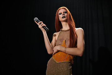 A drag queen in a glamorous outfit holds a microphone, performing on a dark stage.