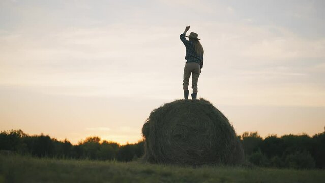 Happy lady dancing on haystack in sunset, merry summer in countryside, back view . Female farmer has fun and joy in early morning in farmland, young woman in country style clothes and hat, good mood