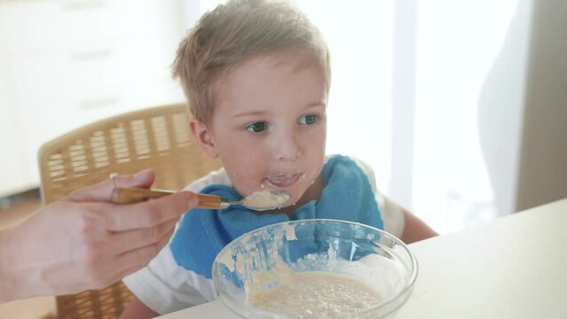 mother feeds porridge to her son. happy family healthy food concept. the boy little son eats porridge with a spoon soiled in his face. child dirty dirty funny lifestyle eating porridge himself video
