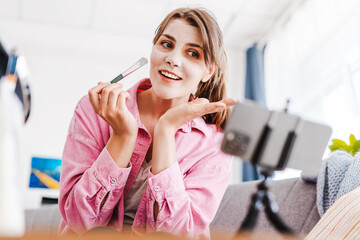 Cheerful woman applying mask on her face with brush, blogger recording video, using mobile phone