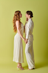 A lesbian couple in white attire stands close together on a green background during their wedding...