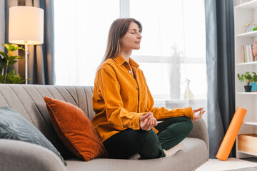Smiling woman sitting on comfortable sofa in lotus position with closed eyes meditating at home