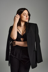 A beautiful plus size woman dressed in a black suit strikes a confident pose against a gray backdrop.