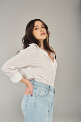 A beautiful plus size woman poses confidently in a white shirt and blue jeans against a gray backdrop.