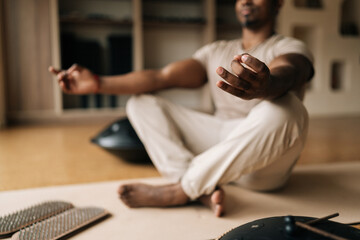 Cropped shot of unrecognizable African young man in trance meditating in lotus pose at home sitting on yoga mat by sadhu board and glucophone. Concept of mindful meditation, wellbeing, mental health.