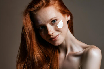 A close-up portrait of a long-haired, smiling young woman with red hair and a bare shoulder. On the cheek there is a smear of cream. Skincare concept