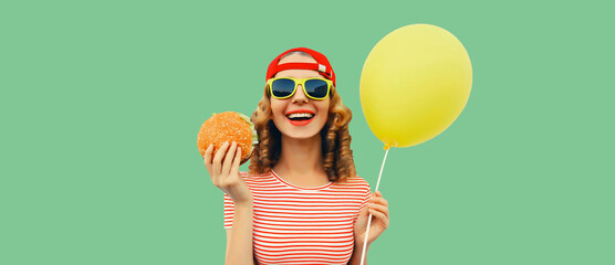 Portrait of cheerful funny young woman with burger fast food and yellow balloon on green background