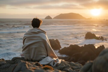 Person on beach rocks, California sunset, oversized blanket, sea waves, golden hour.