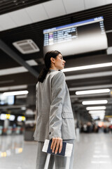 Girl tourist with suitcase looking at flight timetable. Asian woman looks at the flight schedule in the airport terminal to find out information about departure. View from back.