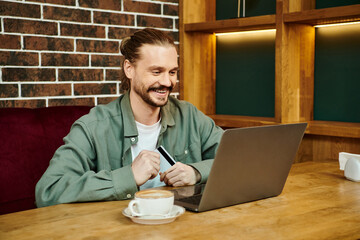 A man sits in a modern cafe, focused on his laptop computer screen, engaged in work or leisure activities.