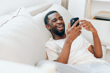 Happy African American man holding a black mobile phone, sitting on a modern sofa in his apartment He is smiling while typing a message and using the internet, enjoying the comfort of his home With a