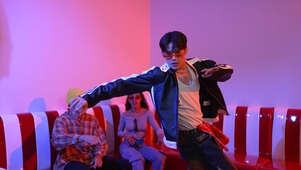 Asian man standing and dancing with pop music while multicultural friend sitting behind at home with led light. Attractive street dancer moving to hip hop music while looking at camera. Regalement.