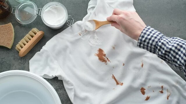 Woman's hand using baking soda powder for eco-friendly stain removal. Splashing cosmetic cream or foundation on a white t-shirt. Zero waste concept. top view