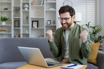 A happy young man sits at home on the sofa in front of a laptop and rejoices in success, showing a...