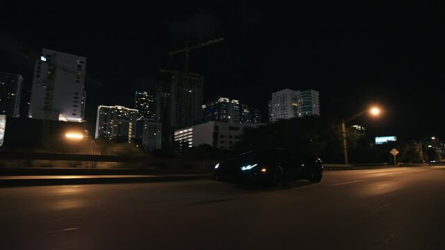Rolling shot of  black supercar driving at night on the streets in Miami Florida city with skyline city lights view