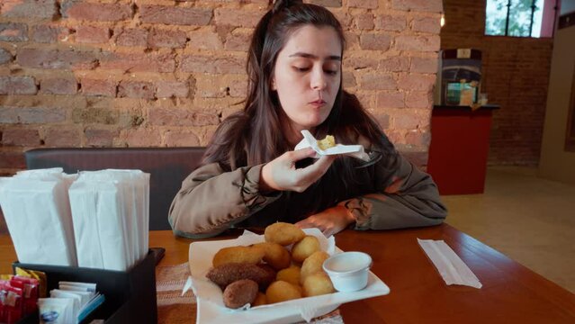 Happy Young Woman Eating a Traditional Brazilian Snack in a Cozy Caf&eacute; Setting. Girl Enjoying Brazilian Fried Snack in Caf&eacute;.