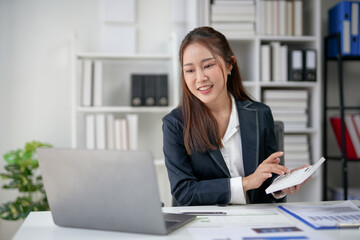 Businesswoman working on a laptop in a modern office, holding a notepad, and smiling. Professional work environment with shelves and documents.
