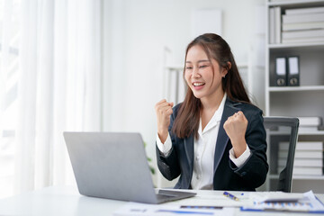 Happy businesswoman celebrating success while working on a laptop in a modern office setting with documents.