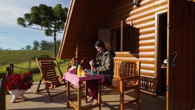 Happy Girl Enjoying Breakfast in a Rustic Cabin. Smiling Young Woman Preparing Breakfast on the Porch of a Wooden Cabin in a Rural Setting