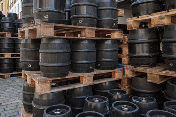 A collection of black barrels neatly stacked on wooden pallets in an outdoor setting. The barrels, possibly used for brewing beer or storage, are arranged in a double-tier formation on the street.