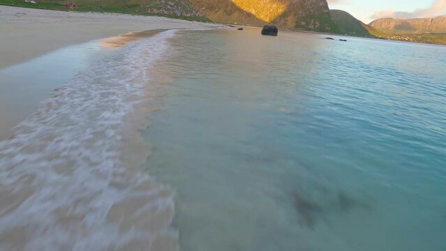 fpv drone flying low and fast over beach and turquoise sea with midnight sun sunset with mountains in the background in Haukland beach Lofoten
