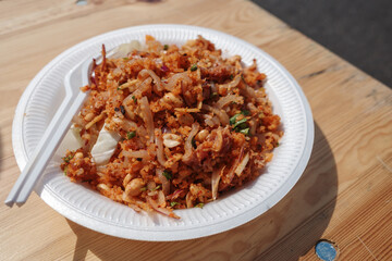 A close-up shot of a delicious plate of spicy Asian street food, Nam khao, served in a white disposable plate on a wooden table.