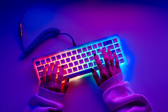 Hands typing on a gaming keyboard with RGB lighting, top view. Female hands typing on colorful gaming keyboard in neon light.
