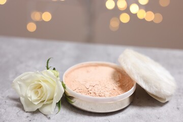 Face powder, puff applicator and rose flower on grey textured table against blurred lights, closeup