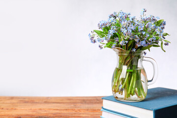 Bouquet of beautiful forget-me-not flowers in glass jug and books on wooden table against light background, space for text