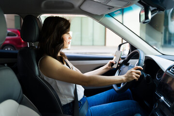 Young woman attentively driving with one hand on the steering wheel and holding a smartphone in the...