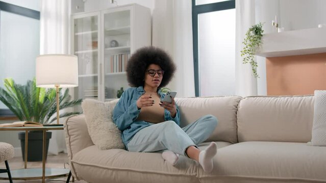 African American woman girl lady user influencer female come in living room walking going in home sit relax at couch sofa using mobile phone internet addict social media smartphone addiction chatting