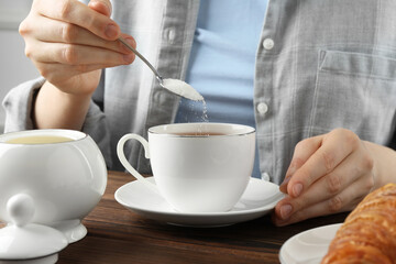 Woman adding sugar into cup of tea at wooden table, closeup