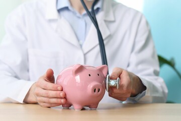 Doctor with piggy bank and stethoscope at wooden table, closeup