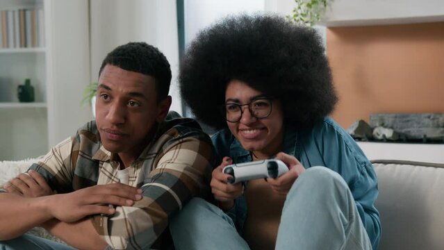 African American couple internet game addict sad offended man guy boyfriend male with happy funny woman female girl girlfriend hold joystick pushing fooling playing console at home fun showing tongue