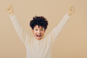 Joyful young boy raising his arms in celebration standing against a soft beige background, exuding...