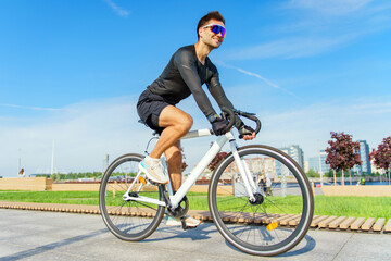 Naklejka premium A man in black cycling attire rides a white bike on a sunny day, smiling, with urban greenery in the background.