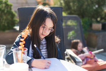 Fototapeta premium A little girl with glasses sits at a table in the backyard and does her homework. Educational concept