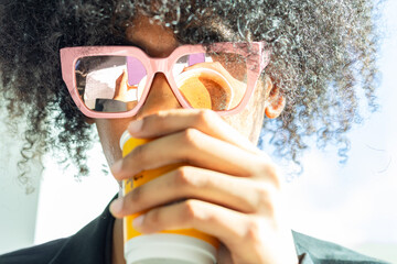 Young mixed-race non-binary person with an afro and sunglasses, wearing a black business suit. Drinking coffee while the reflection is visible in the sunglasses.