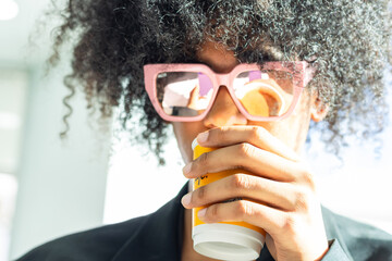 Non-binary person with an afro and sunglasses, wearing a black business suit. Drinking coffee while their reflection is visible in the sunglasses